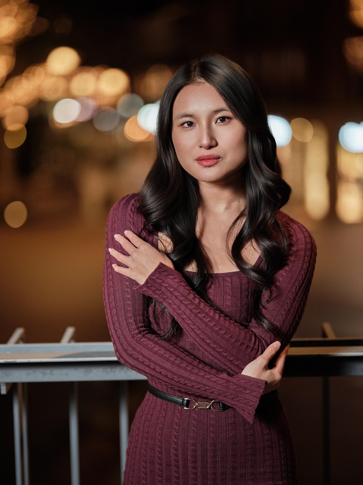 Avondportret van een jonge vrouw in een bordeauxrode jurk met armen over elkaar, staand tegen een railing met zachte stadslichten op de achtergrond.