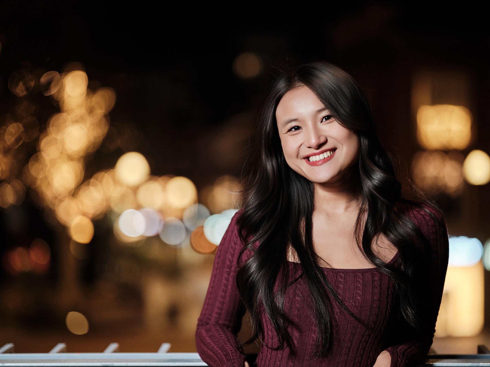 Lachend avondportret van een jonge vrouw in bordeauxrode jurk, leunend op een railing met kleurrijke stadslichten in de bokeh-achtergrond.