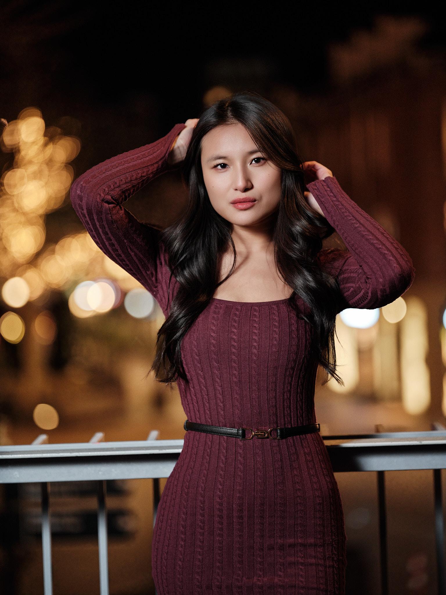 Avondportret van een jonge vrouw in bordeauxrode jurk met de handen in haar haar, met warme stadslichten in de onscherpe achtergrond.