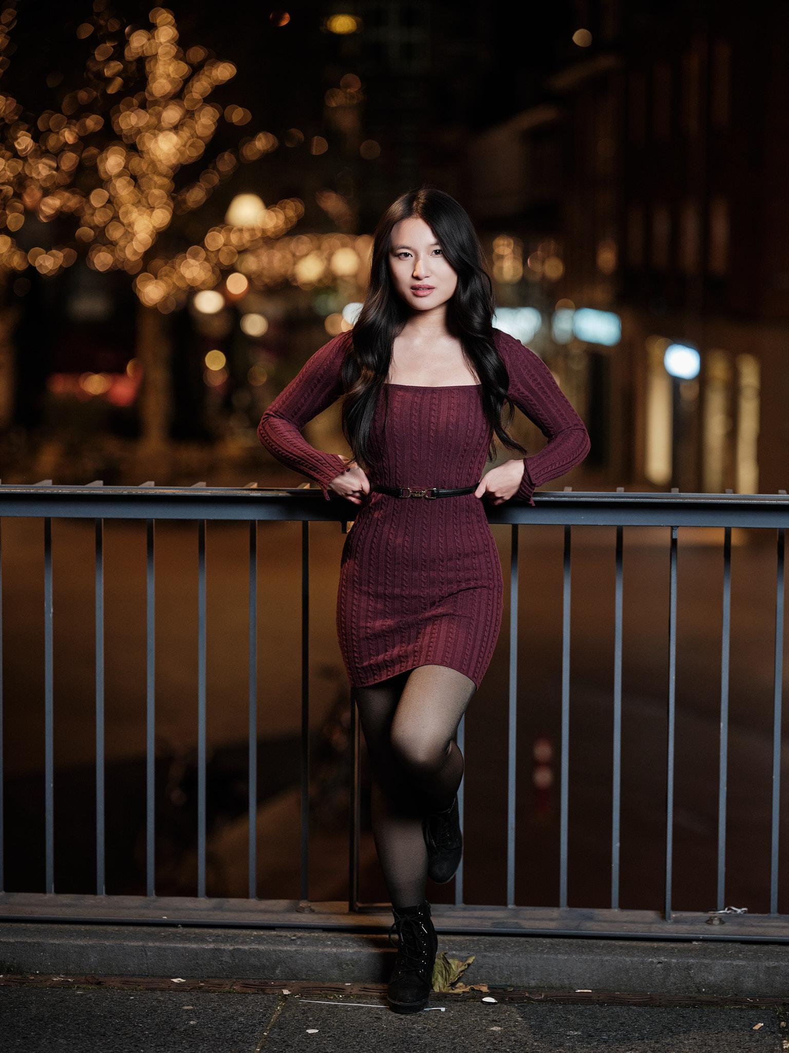 Volledig avondportret van een jonge vrouw in bordeauxrode jurk, leunend tegen een railing met verlichte bomen en stadslichten op de achtergrond.