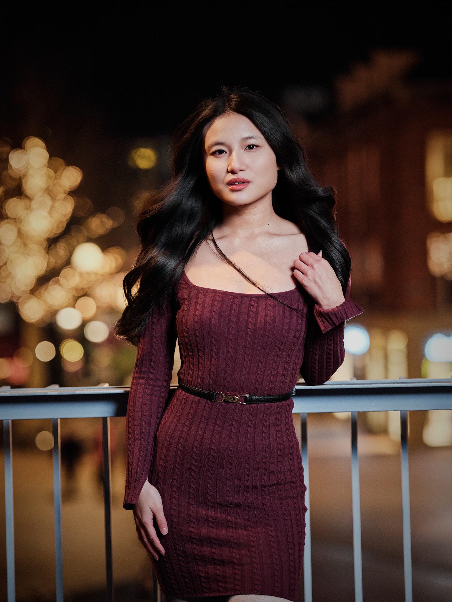 Avondportret van een jonge vrouw in bordeauxrode jurk, leunend tegen een railing met verlichte bomen en zachte bokeh-achtergrond.