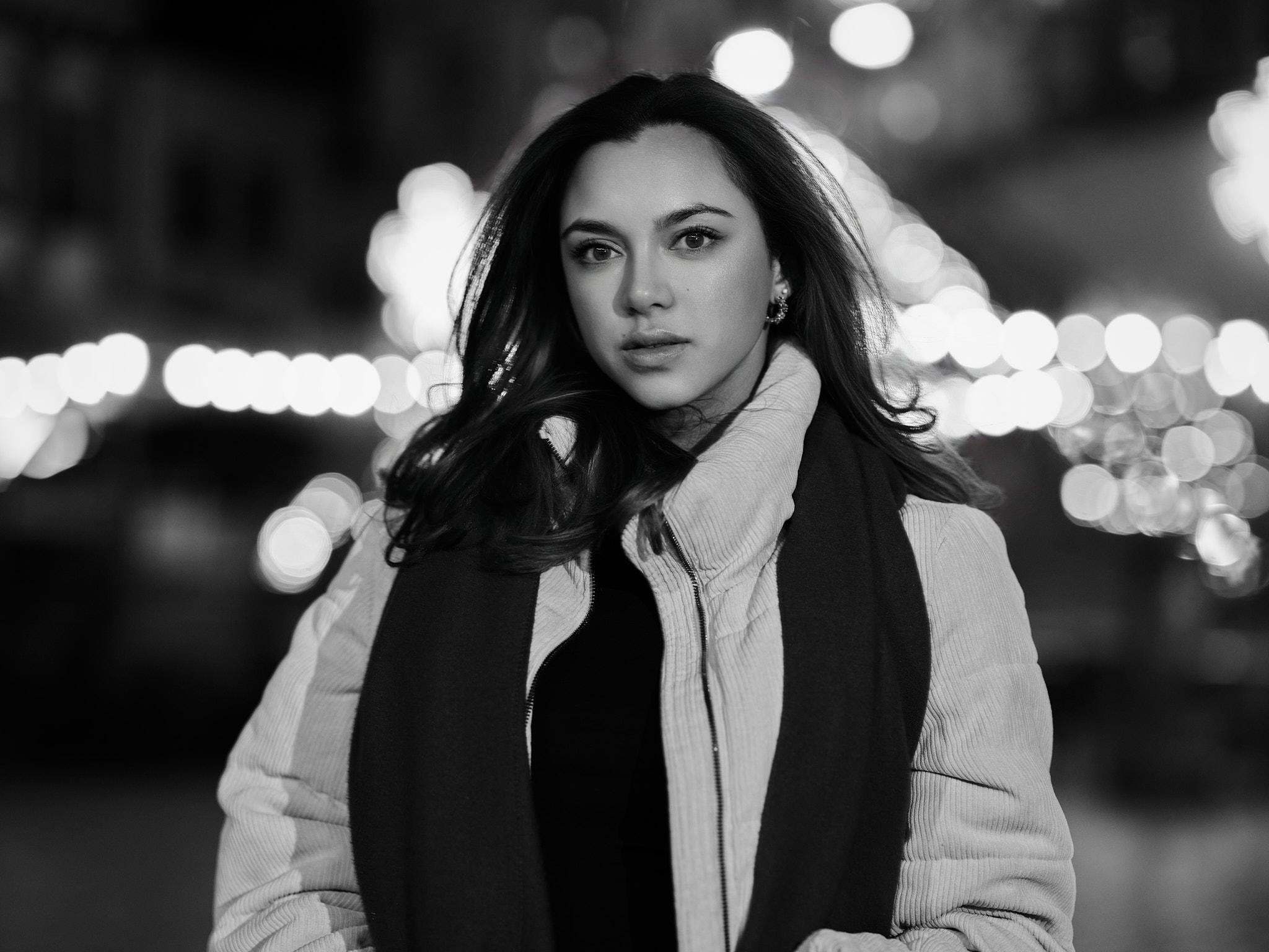 Zwart-wit avondportret van een jonge vrouw in winterjas en sjaal, met zachte lichtcirkels van de stad op de achtergrond.